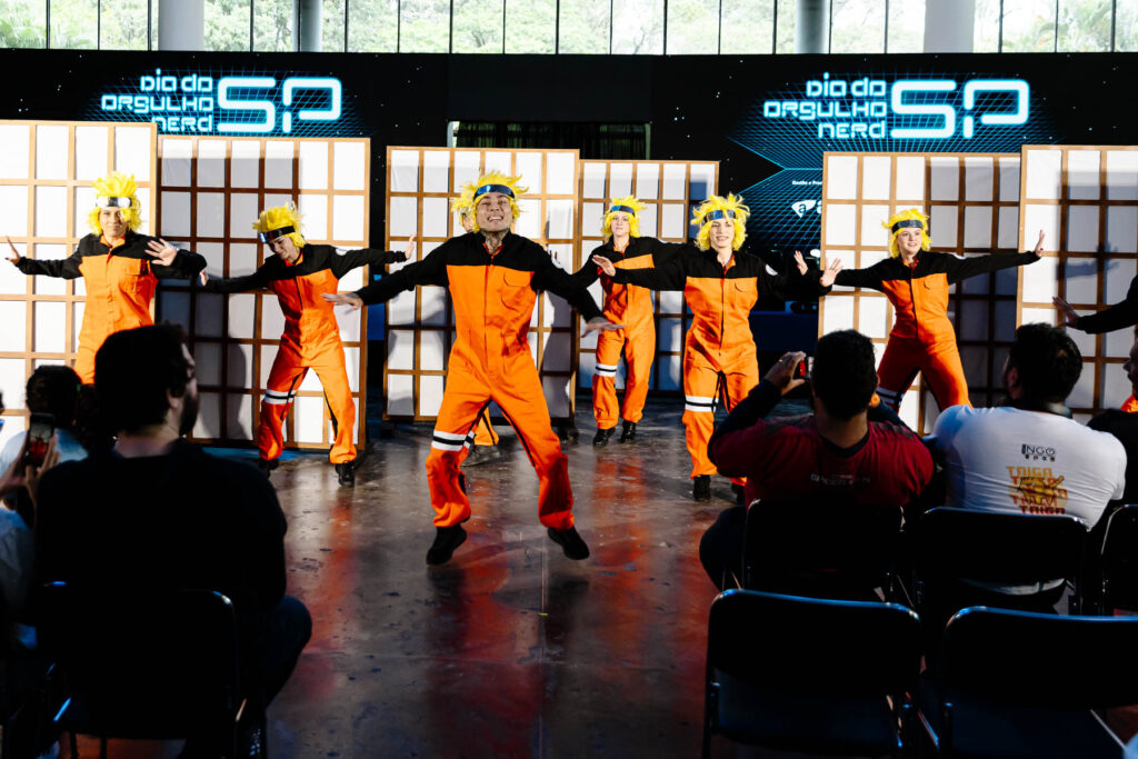 Na imagem, um grupo de seis cosplayers, vestidos como o personagem Naruto Uzumaki, está realizando uma performance. Cada cosplayer usa um macacão laranja e preto, típico do personagem, e tem cabelo amarelo espetado. Eles estão alinhados e executam uma coreografia, com braços estendidos e posturas de ação. Ao fundo, há uma parede decorada com o texto "Dia do Orgulho Nerd" e o logotipo "SP". O ambiente tem um piso de concreto e um grande espaço onde o público sentado e algumas pessoas em pé observam e fotografam a apresentação.